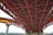 A detailed view of a large steel bridge structure with red and orange iron beams intersecting in a geometric pattern. The bridge is elevated, supported by concrete pillars, with a cityscape visible in the background.