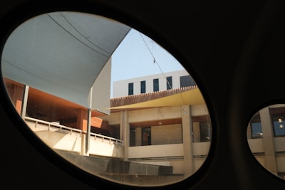 The architectural scene is captured through large, circular windows framing a courtyard. The central structure features concrete surfaces and glass windows, topped with metal panels and an overhead shade. The view presents a mix of geometric lines and surfaces, creating a modern urban aesthetic.