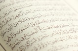 Arabic script with ornate, artistic calligraphy on the pages of an open book. The text is precise with diacritical marks, set against an off-white background.