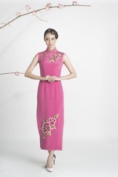 A person wears a bright pink, traditional-style dress adorned with floral embroidery. The dress is sleeveless and floor-length with intricate floral designs near the neckline and bottom. The individual is standing on a white background, with their hair styled elegantly. A branch with delicate pink blossoms extends across the top of the scene, adding a touch of nature.