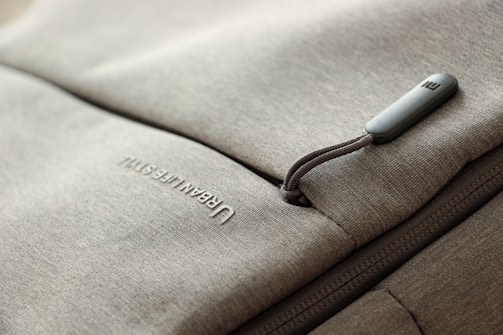 A close-up of a grey fabric bag with a zippered pocket. The word 'URBAN LIFESTYLE' is printed in white text on the fabric. The zipper pull has a small, rectangular, grey tag attached to it.