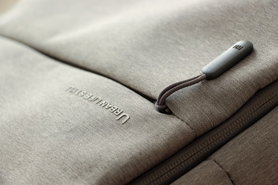 A close-up of a grey fabric bag with a zippered pocket. The word 'URBAN LIFESTYLE' is printed in white text on the fabric. The zipper pull has a small, rectangular, grey tag attached to it.