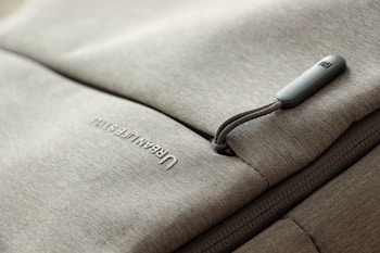 A close-up of a grey fabric bag with a zippered pocket. The word 'URBAN LIFESTYLE' is printed in white text on the fabric. The zipper pull has a small, rectangular, grey tag attached to it.