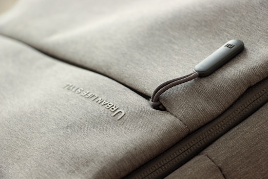 A close-up of a grey fabric bag with a zippered pocket. The word 'URBAN LIFESTYLE' is printed in white text on the fabric. The zipper pull has a small, rectangular, grey tag attached to it.