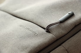 A close-up of a grey fabric bag with a zippered pocket. The word 'URBAN LIFESTYLE' is printed in white text on the fabric. The zipper pull has a small, rectangular, grey tag attached to it.