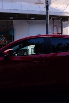 A red car passes by in front of a commercial building with white walls and a dark overhang. There is a visible support structure and a bit of signage. The car's windows reflect some light, and there are slight shadows and reflections on the vehicle.