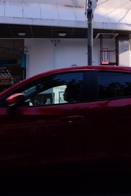 A red car passes by in front of a commercial building with white walls and a dark overhang. There is a visible support structure and a bit of signage. The car's windows reflect some light, and there are slight shadows and reflections on the vehicle.