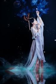 A woman dressed in flowing, ethereal robes reaches up towards glowing blue branches set against a dark, starry background. She wears antler-like adornments on her head and her reflection is visible in the water below, surrounded by mist.