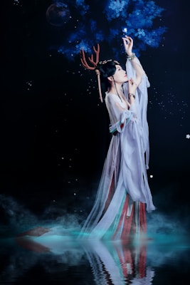 A woman dressed in flowing, ethereal robes reaches up towards glowing blue branches set against a dark, starry background. She wears antler-like adornments on her head and her reflection is visible in the water below, surrounded by mist.