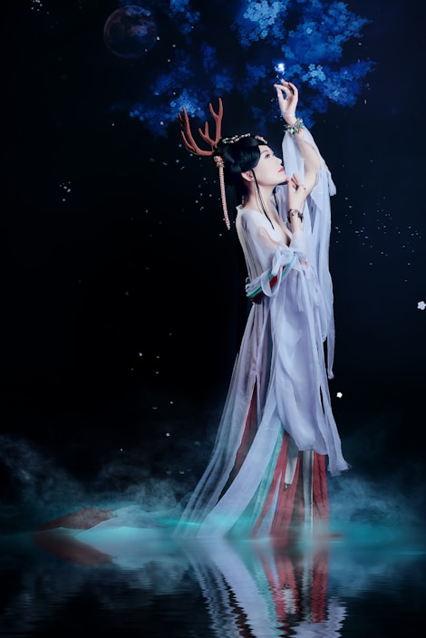 A woman dressed in flowing, ethereal robes reaches up towards glowing blue branches set against a dark, starry background. She wears antler-like adornments on her head and her reflection is visible in the water below, surrounded by mist.