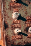 A close-up of a weathered metal control panel featuring toggle switches and embossed labels for operations such as 'LOWER', 'IN', 'OUT', 'OPEN', and 'CLOSE'. The panel is rustic with signs of rust and wear, suggesting age and outdoor exposure.