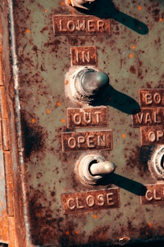 A close-up of a weathered metal control panel featuring toggle switches and embossed labels for operations such as 'LOWER', 'IN', 'OUT', 'OPEN', and 'CLOSE'. The panel is rustic with signs of rust and wear, suggesting age and outdoor exposure.