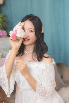 A woman wearing an off-shoulder, lace-patterned white dress is holding a small plush toy decorated with a pink tassel in front of one eye. Her hair is dark and styled with a pearl hair accessory. She has a playful and cheerful expression.