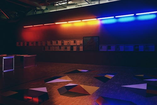 A dimly lit room with modern art installations features a wall lined with framed artworks under a row of vivid, multicolored fluorescent lights. The floor displays geometric shapes in dark hues.
