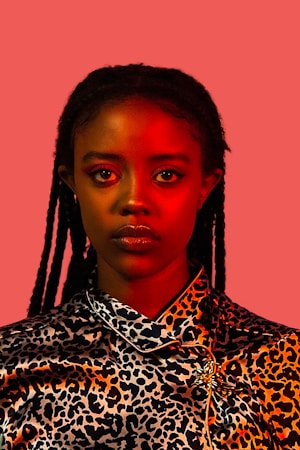 A person with braided hair appears against a solid, muted red background. The upper part of their body is visible, wearing a leopard print garment with a decorative brooch. The lighting casts a warm, reddish tone across their face, adding depth and contrast to the overall composition.