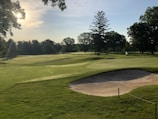 A golf course with well-manicured greens and sand traps, surrounded by lush trees. The sun casts a warm glow over the landscape, creating elongated shadows. The area is peaceful and serene, with the flag indicating a hole on the green.