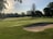 A golf course with well-manicured greens and sand traps, surrounded by lush trees. The sun casts a warm glow over the landscape, creating elongated shadows. The area is peaceful and serene, with the flag indicating a hole on the green.