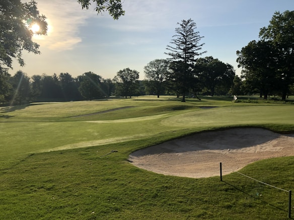 A golf course with well-manicured greens and sand traps, surrounded by lush trees. The sun casts a warm glow over the landscape, creating elongated shadows. The area is peaceful and serene, with the flag indicating a hole on the green.