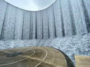 A large, curved wall features cascading water creating a waterfall effect. The ground shows wet patterns from the falling water, giving a sense of movement and fluidity. The sky above is overcast, adding a serene and calm atmosphere.