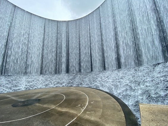 A large, curved wall features cascading water creating a waterfall effect. The ground shows wet patterns from the falling water, giving a sense of movement and fluidity. The sky above is overcast, adding a serene and calm atmosphere.