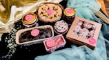An arrangement of various dessert items including a pie, cupcakes, cake slices, and jarred goods. Each item is decorated and labeled with 'Sugarb'. The background is composed of draped fabric, adding an elegant touch.