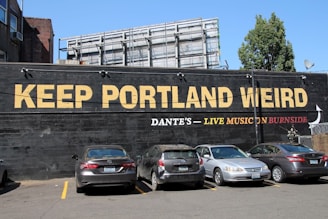 A large mural painted on the side of a building, featuring bold yellow and red lettering. Below the text, there is a line for parking where several cars are parked facing the mural. The scene includes a tree and a billboard structure above the mural, with a clear blue sky overhead.