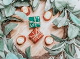 A decorative arrangement consists of small wrapped gifts placed on a wooden surface, surrounded by green leaves and small candles. The gifts are wrapped in shiny red and green paper with golden ribbons. The candles are positioned in orange holders, adding a warm ambiance.