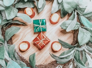 A decorative arrangement consists of small wrapped gifts placed on a wooden surface, surrounded by green leaves and small candles. The gifts are wrapped in shiny red and green paper with golden ribbons. The candles are positioned in orange holders, adding a warm ambiance.