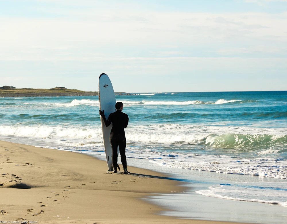 A surfer in a wetsuit standing at the water's edge with a board