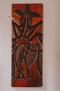 A carved wooden artwork depicts a figure holding a large fish, with a palm tree, mountains, and a small house in the background. The carving features thick, detailed lines and predominantly dark brown tones with a glossy finish, creating a rustic and traditional aesthetic.