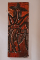 A carved wooden artwork depicts a figure holding a large fish, with a palm tree, mountains, and a small house in the background. The carving features thick, detailed lines and predominantly dark brown tones with a glossy finish, creating a rustic and traditional aesthetic.