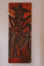 A carved wooden artwork depicts a figure holding a large fish, with a palm tree, mountains, and a small house in the background. The carving features thick, detailed lines and predominantly dark brown tones with a glossy finish, creating a rustic and traditional aesthetic.