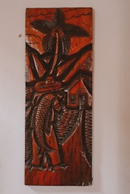 A carved wooden artwork depicts a figure holding a large fish, with a palm tree, mountains, and a small house in the background. The carving features thick, detailed lines and predominantly dark brown tones with a glossy finish, creating a rustic and traditional aesthetic.