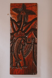 A carved wooden artwork depicts a figure holding a large fish, with a palm tree, mountains, and a small house in the background. The carving features thick, detailed lines and predominantly dark brown tones with a glossy finish, creating a rustic and traditional aesthetic.