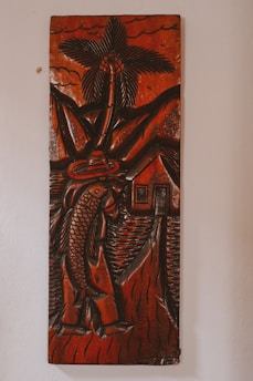 A carved wooden artwork depicts a figure holding a large fish, with a palm tree, mountains, and a small house in the background. The carving features thick, detailed lines and predominantly dark brown tones with a glossy finish, creating a rustic and traditional aesthetic.