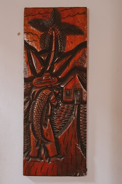 A carved wooden artwork depicts a figure holding a large fish, with a palm tree, mountains, and a small house in the background. The carving features thick, detailed lines and predominantly dark brown tones with a glossy finish, creating a rustic and traditional aesthetic.