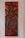 A carved wooden artwork depicts a figure holding a large fish, with a palm tree, mountains, and a small house in the background. The carving features thick, detailed lines and predominantly dark brown tones with a glossy finish, creating a rustic and traditional aesthetic.