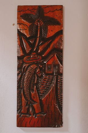 A carved wooden artwork depicts a figure holding a large fish, with a palm tree, mountains, and a small house in the background. The carving features thick, detailed lines and predominantly dark brown tones with a glossy finish, creating a rustic and traditional aesthetic.