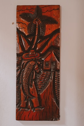 A carved wooden artwork depicts a figure holding a large fish, with a palm tree, mountains, and a small house in the background. The carving features thick, detailed lines and predominantly dark brown tones with a glossy finish, creating a rustic and traditional aesthetic.
