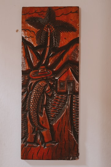 A carved wooden artwork depicts a figure holding a large fish, with a palm tree, mountains, and a small house in the background. The carving features thick, detailed lines and predominantly dark brown tones with a glossy finish, creating a rustic and traditional aesthetic.