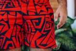 Bright red shorts with bold, abstract black patterns are prominently displayed. A person’s hand is partially visible on the right side, and there are green leaves blurred in the background.