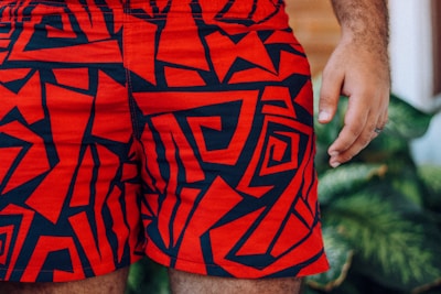 Bright red shorts with bold, abstract black patterns are prominently displayed. A person’s hand is partially visible on the right side, and there are green leaves blurred in the background.