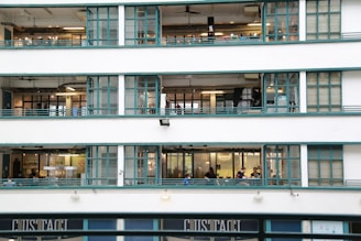 A multi-story building with large glass windows, featuring teal metal frames. Inside, people are visible engaging in various activities on different floors. The building exudes an industrial and functional architecture style.