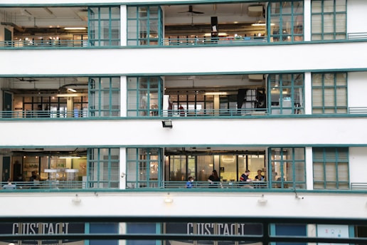 A multi-story building with large glass windows, featuring teal metal frames. Inside, people are visible engaging in various activities on different floors. The building exudes an industrial and functional architecture style.