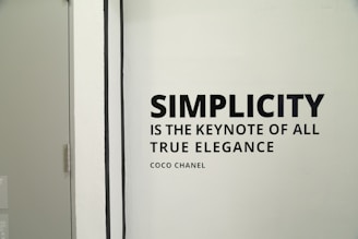 A monochrome wall features the quote 'Simplicity is the keynote of all true elegance' by Coco Chanel. The quote is in bold, black font on a light background. To the left side of the image, a simple door with a metal handle is partially visible.