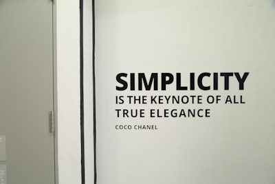 A monochrome wall features the quote 'Simplicity is the keynote of all true elegance' by Coco Chanel. The quote is in bold, black font on a light background. To the left side of the image, a simple door with a metal handle is partially visible.