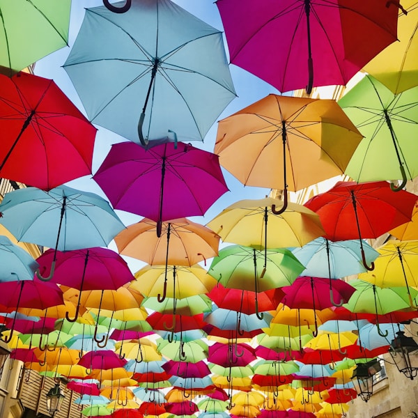 A vibrant display of numerous colorful umbrellas suspended in the air, forming a canopy with a clear blue sky peeking through. The image showcases a playful and whimsical arrangement, with the umbrellas overlapping in various shades including pink, red, orange, yellow, green, and blue.