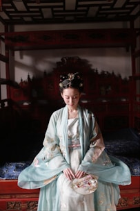 A woman in traditional clothing sits serenely on a carved wooden bed, gently holding a decorative fan. Her ornate robe features delicate patterns in soft pastel colors, and her hair is elegantly styled and adorned with intricate hairpieces. The room has an old-world charm, with rich wooden textures and intricate details.