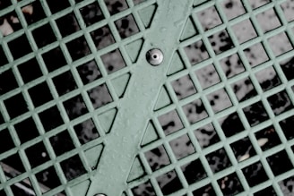 A close-up view of a mint green metal grid with intersecting lines forming squares, featuring droplets of water on the surface. A bolt is visible at the center of the diagonal support bar running through the grid, with a dark background visible through the open spaces between the metal.