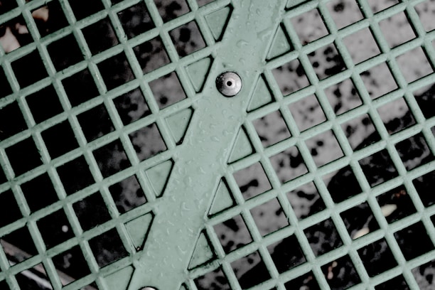 A close-up view of a mint green metal grid with intersecting lines forming squares, featuring droplets of water on the surface. A bolt is visible at the center of the diagonal support bar running through the grid, with a dark background visible through the open spaces between the metal.